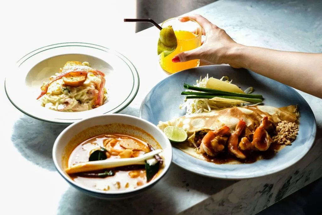 Fine dining meal set in a marble table, consisting of pasta, soup, vegetables, seafood, and a colorful tropical drink garnished with fruit.