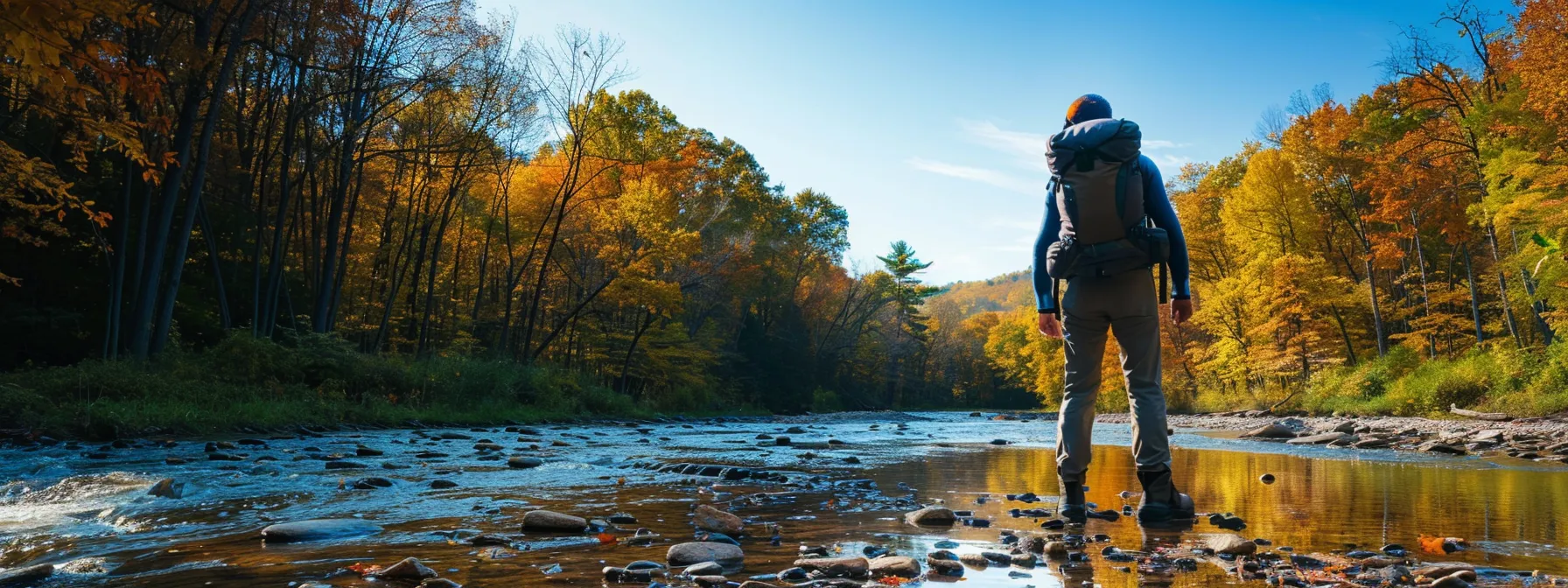a hiker with a backpack standing by a tranquil river in cuyahoga valley national park, surrounded by lush oak forests under a clear blue sky.